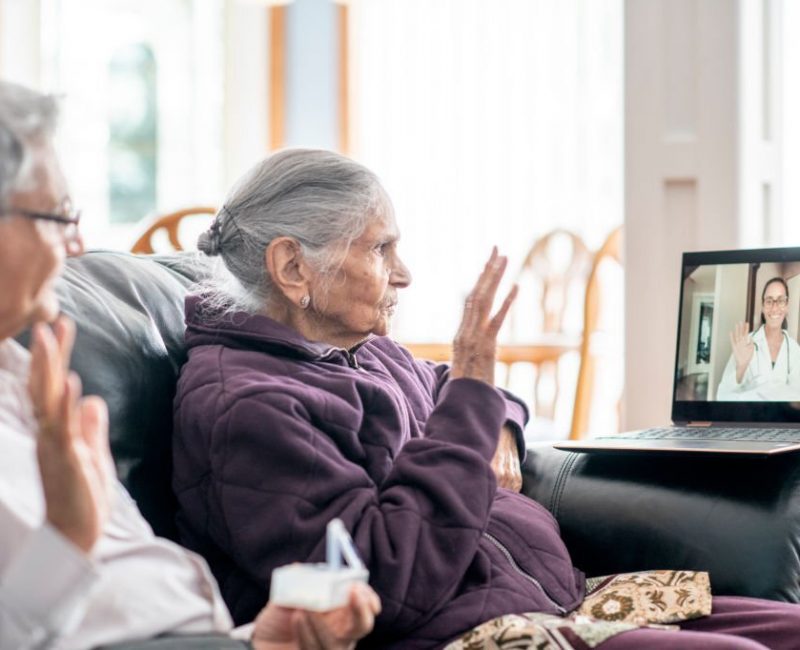 An elderly woman of Indian ethnicity sits on the couch and video chats with a doctor during a virtual appointment. Her family member sits next to her.