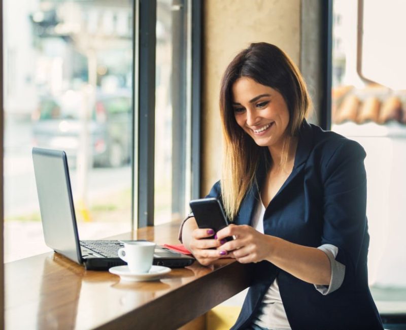 Businesswoman Using Phone Working On Laptop In Coffee Shop